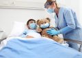 nurse takes care of the patient child in hospital bed playing with teddy bear, wearing protective masks, corona virus covid 19 protection concept,