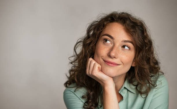 Portrait of young thoughtful woman with hand on chin having an idea against grey background. Beautiful pensive woman looking away while thinking. Close up face of natural girl planning her future isolated on gray wall with copy space.