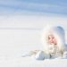Beautiful baby in a white suit sitting in a snow field on a very sunny winter day