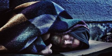 A homeless man sleeps under a ratty blanket on a hard stone sidewalk