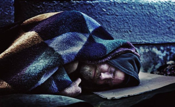 A homeless man sleeps under a ratty blanket on a hard stone sidewalk